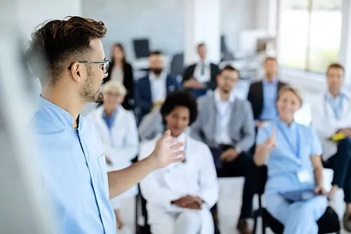 A man in a light blue shirt, with short brown hair and glasses, speaks to a diverse group of people seated in a room. The audience, dressed in a mix of casual and professional attire, appears attentive and engaged.