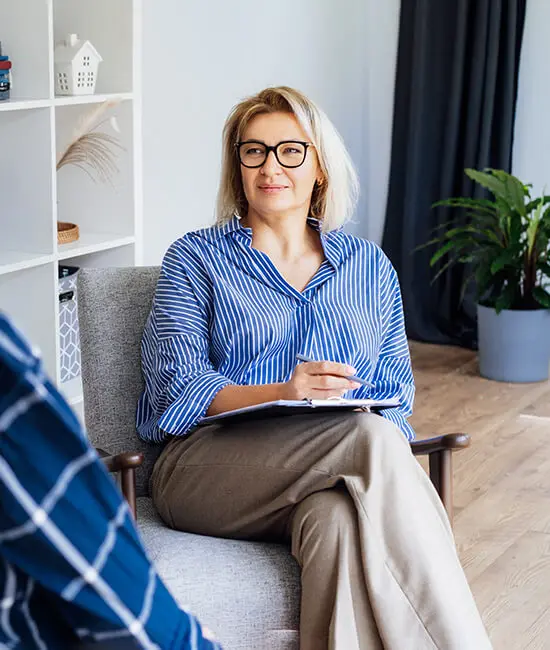 A person with blonde hair and glasses sits on a chair, smiling and holding a notebook with a pen. They wear a blue and white striped shirt and beige pants. A plant and a bookshelf are in the background.