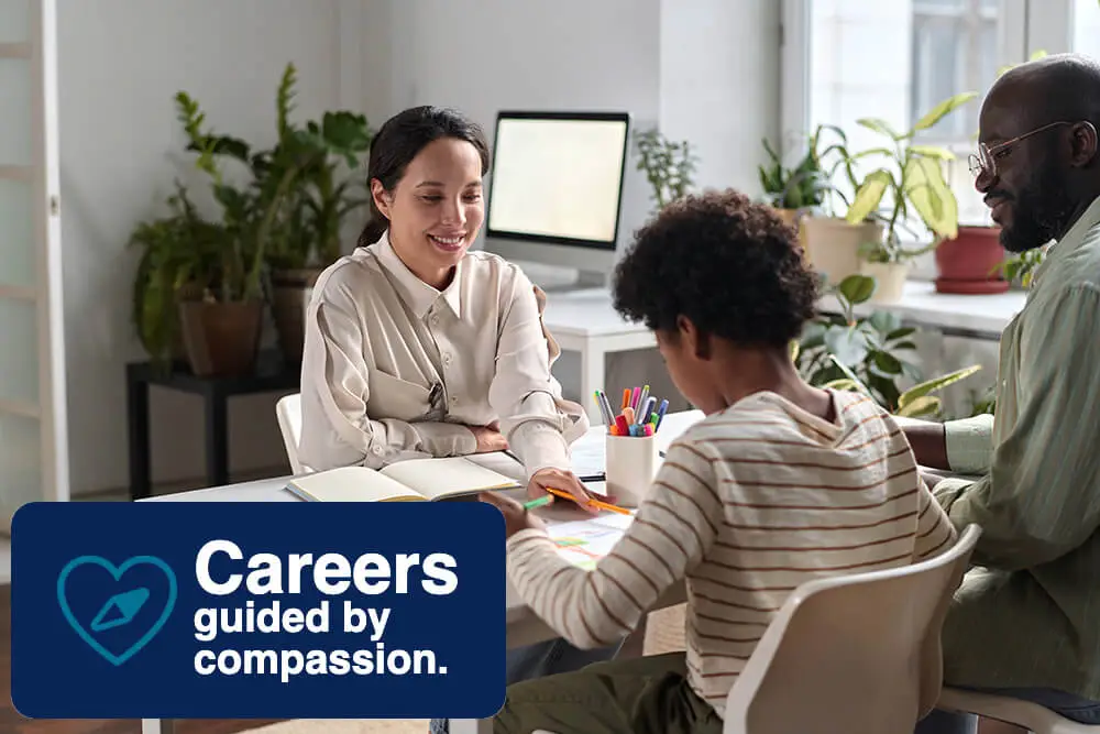 A woman is sitting at a desk with a young boy and a man. They are engaged in a discussion. The room has plants and a computer. A graphic reads Careers guided by compassion with a heart and pen symbol.