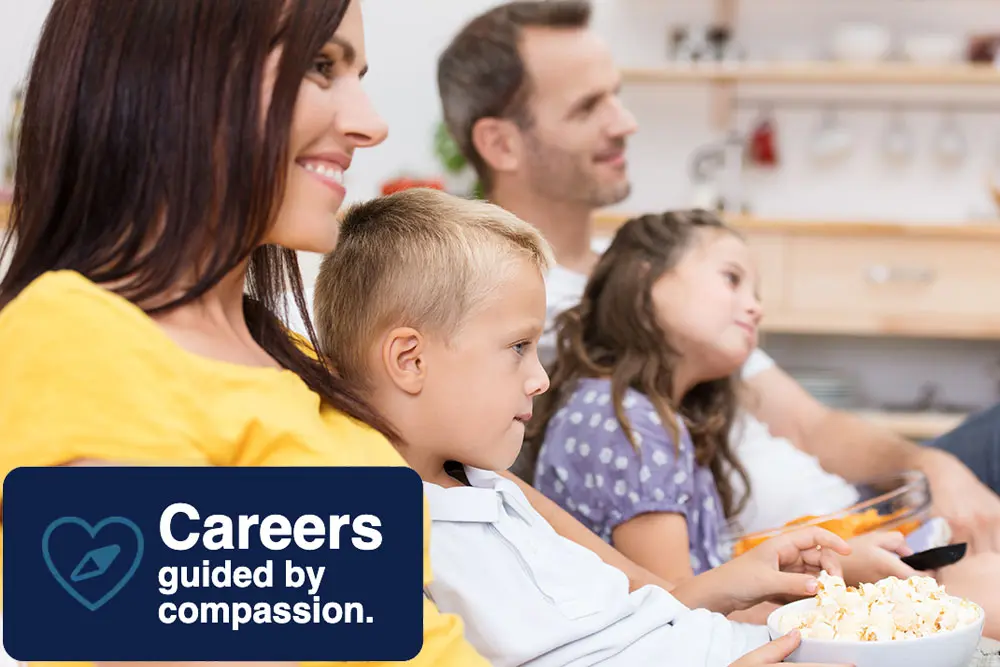 A family of four, two adults and two children, sit closely on a couch smiling and watching something off-camera. A dark blue rectangle with a heart icon contains the text Careers guided by compassion.