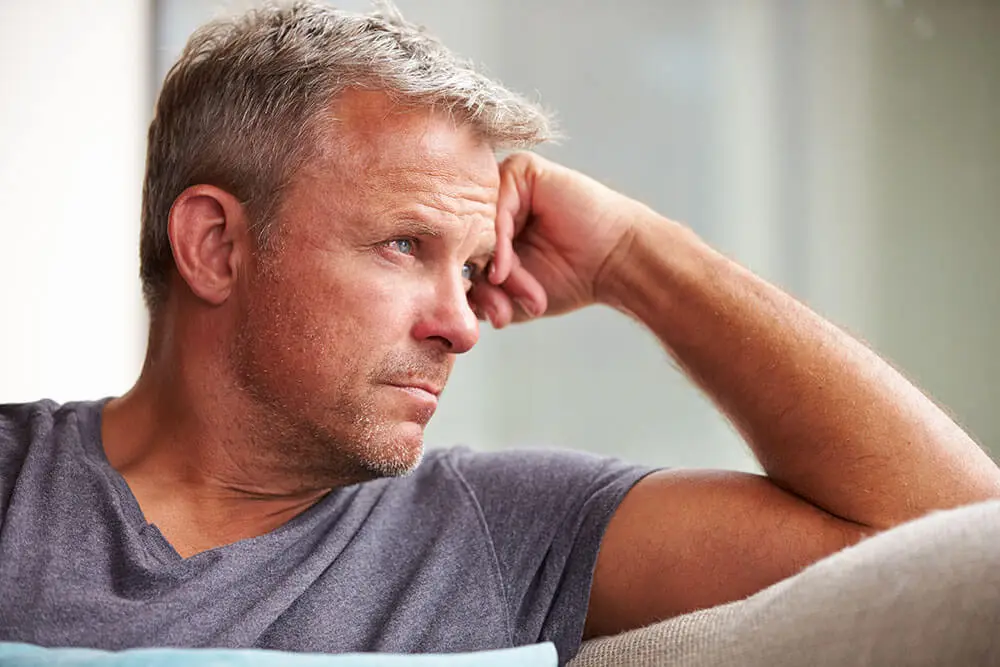 Man with short gray hair and stubble, wearing a gray T-shirt, gazes intently into the distance while resting his head on his hand. He is seated indoors, with a blurred background.