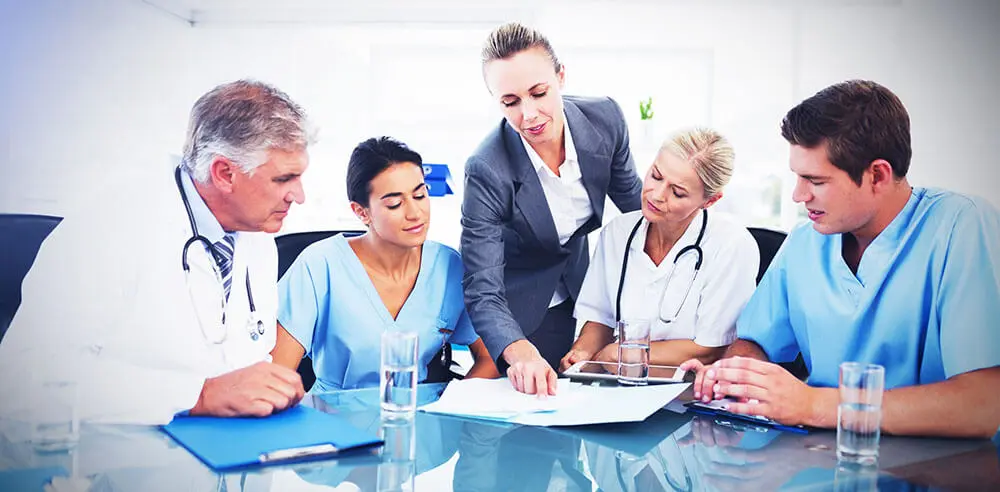 A group of medical professionals and a businessperson in a meeting room. Two are in white coats, three in blue scrubs. The businessperson, in a gray suit, points at documents on the table. They are discussing something intently.