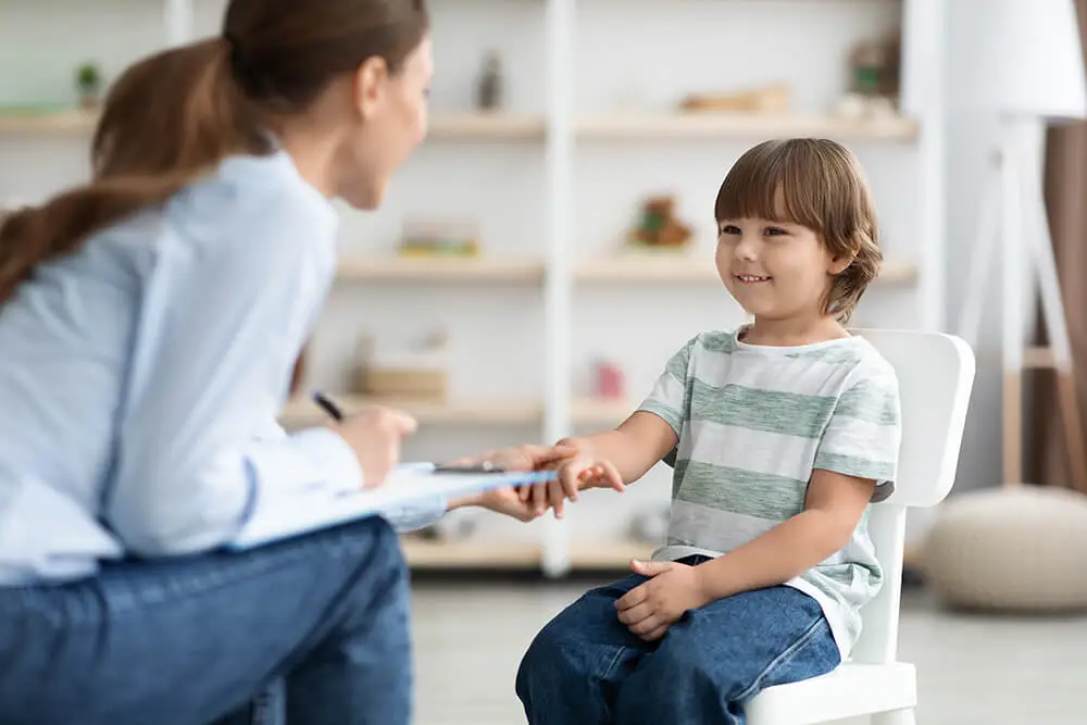 A woman sits talking to a young boy who is smiling and seated on a chair. They are in a bright room with shelves in the background. The woman holds a clipboard, possibly engaging with the child in a friendly manner.