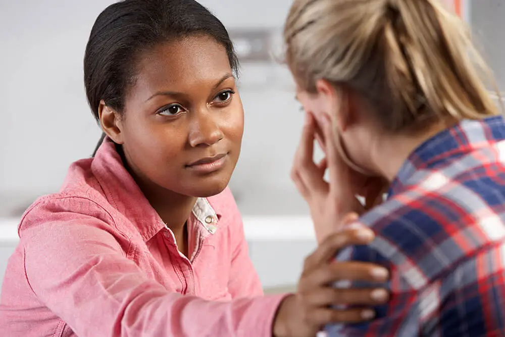 A woman in a pink shirt is comforting another woman in a plaid shirt by gently touching her shoulder. The second woman appears to be upset, covering part of her face with her hand.