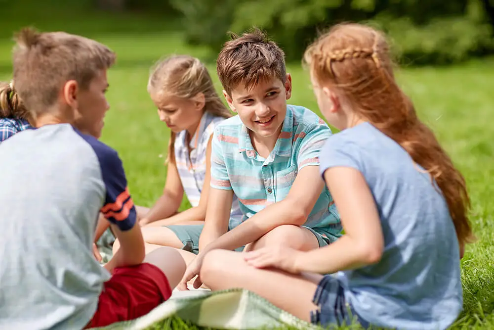 A group of four children sit on the grass talking. They are casually dressed and facing each other, with two boys and two girls. Trees and grass are visible in the background, suggesting a park or garden setting.
